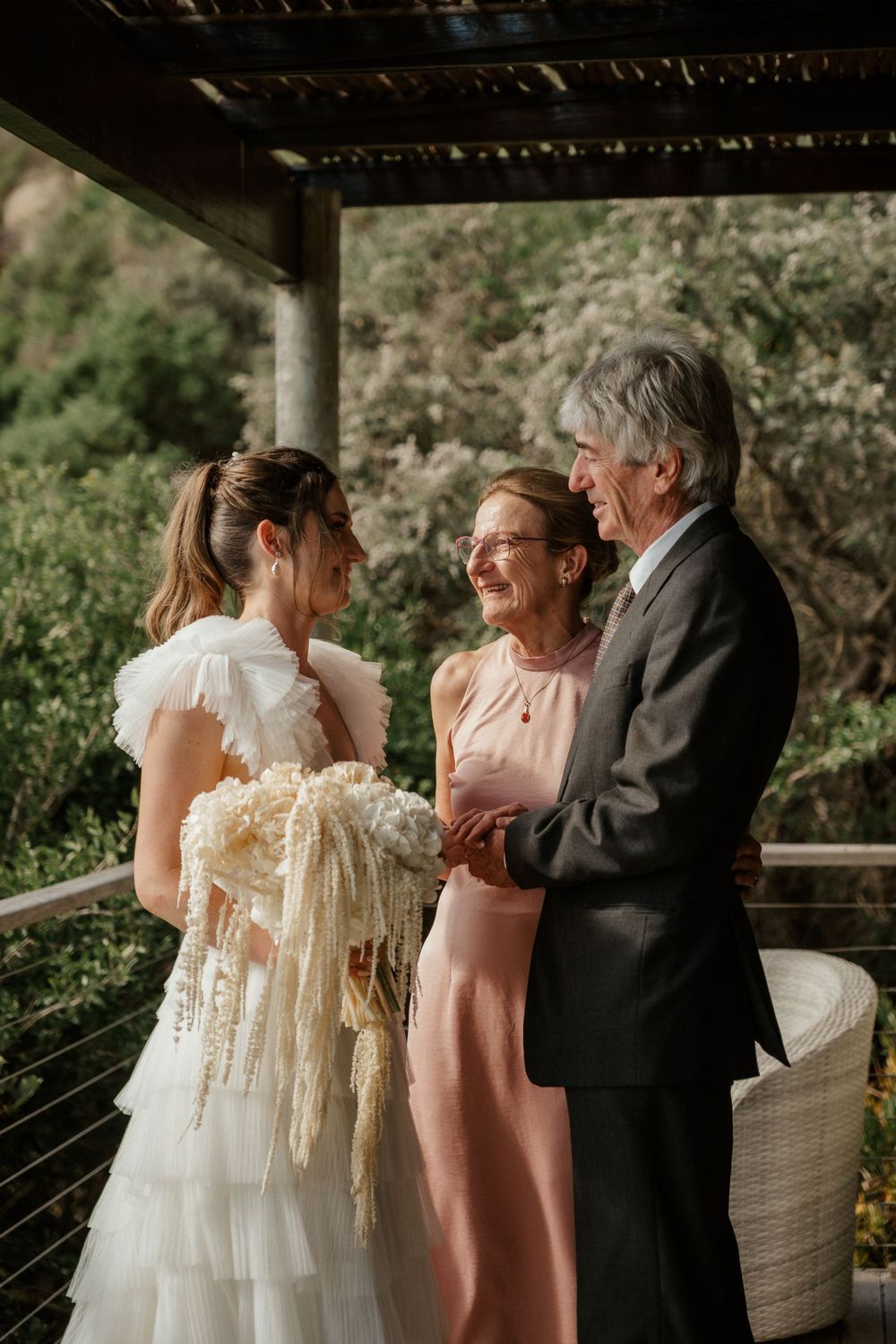 Three people share an emotional moment during an outdoor wedding ceremony under a covered porch.