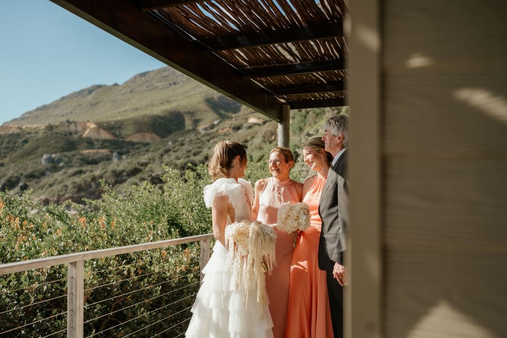 Wedding party gathers on a scenic terrace overlooking mountain landscape with natural lighting and rustic wooden pergola.