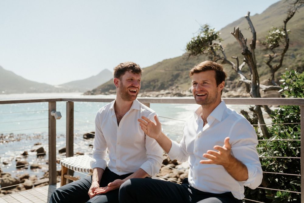 Two people sharing a laugh on a coastal deck with mountains and ocean in the background.