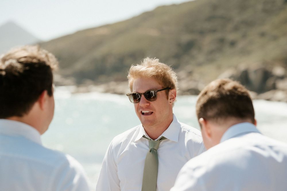 Groomsmen chat by the ocean on a sunny day with coastal mountains in view.