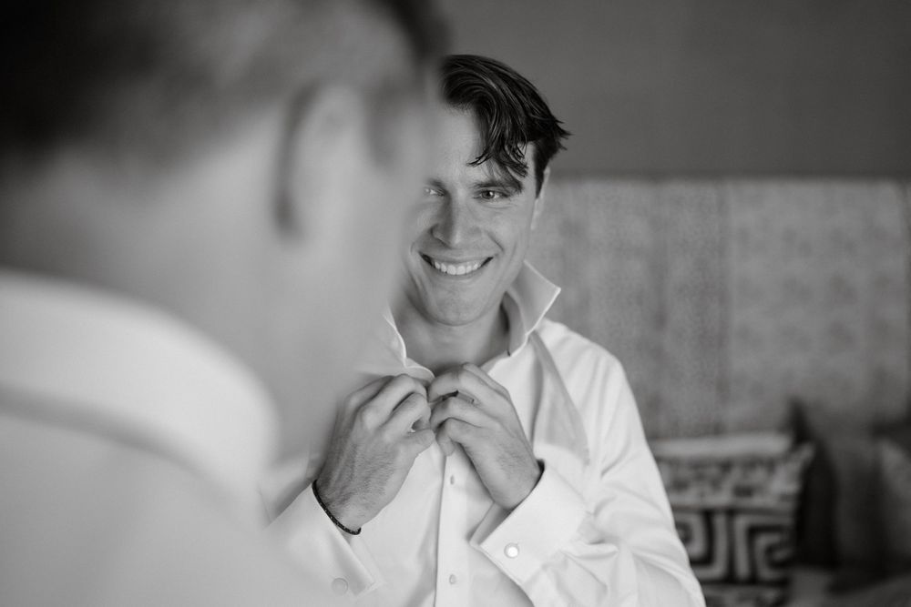 A black and white photograph shows a person wearing a white dress shirt while buttoning up and smiling.
