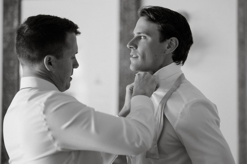 A groomsman helps adjust another's bow tie in an intimate black and white pre-wedding moment.