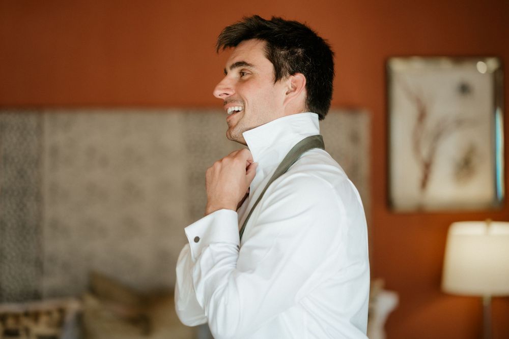 A smiling person in a white dress shirt adjusts their collar while standing in a warmly lit room.