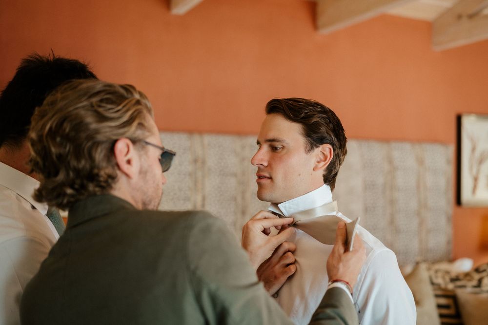 Two people adjusting formal attire during wedding preparation in a coral-colored room.
