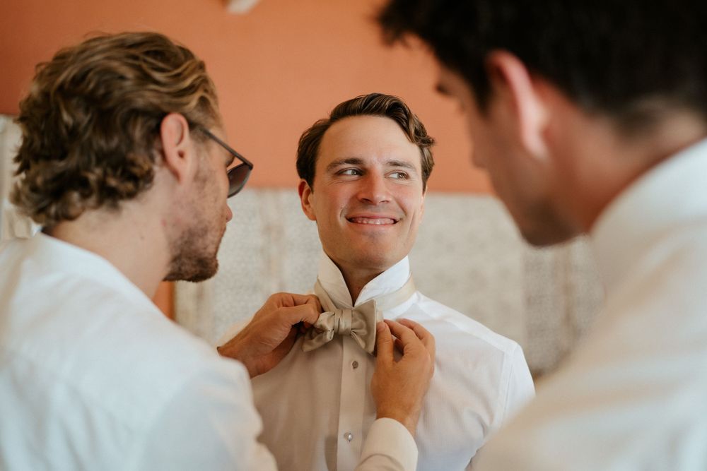 Groomsmen help adjust the bowtie of a smiling groom during wedding preparations.