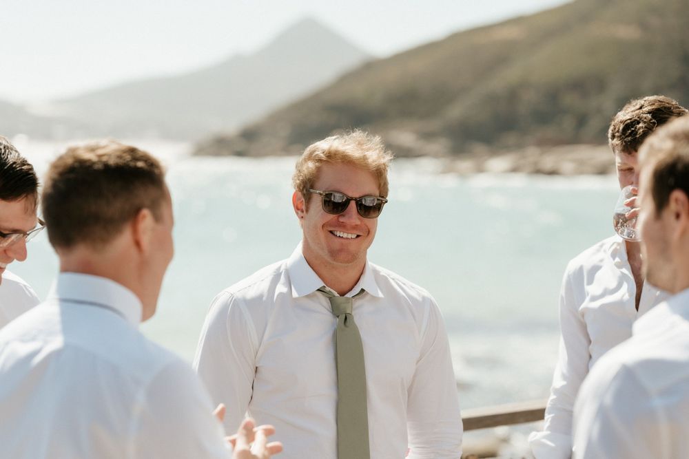 Group of men in white shirts and ties laughing at coastal wedding event.
