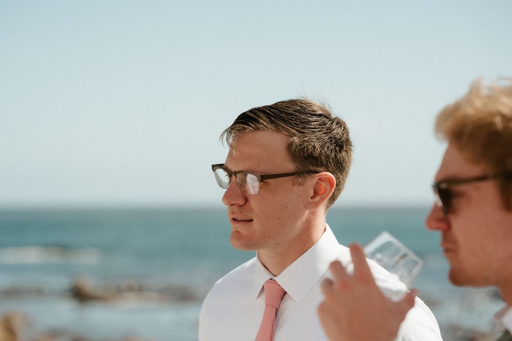 Men in formal attire gathered by the ocean on sunny day.