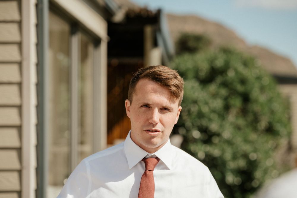 A person in a white dress shirt and orange tie stands outside a house on a sunny day.