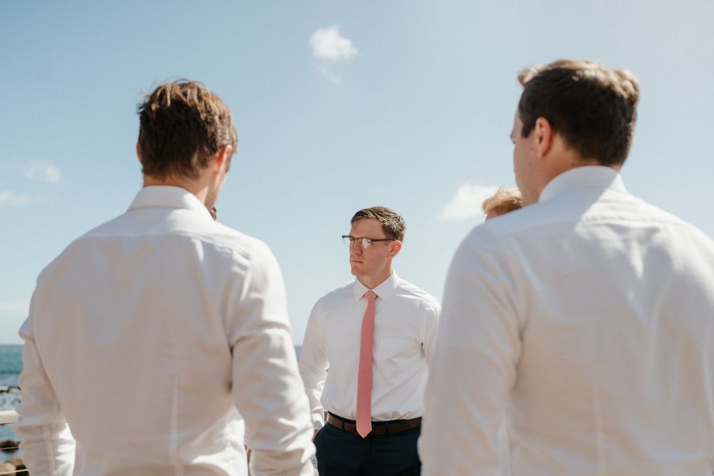 Three people in white and pink formal attire stand together against a bright blue sky.