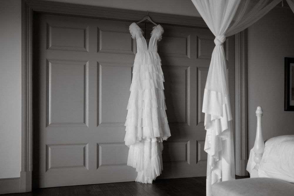 Two elegant white wedding dresses hanging on a gray paneled wall in a soft black and white photograph.