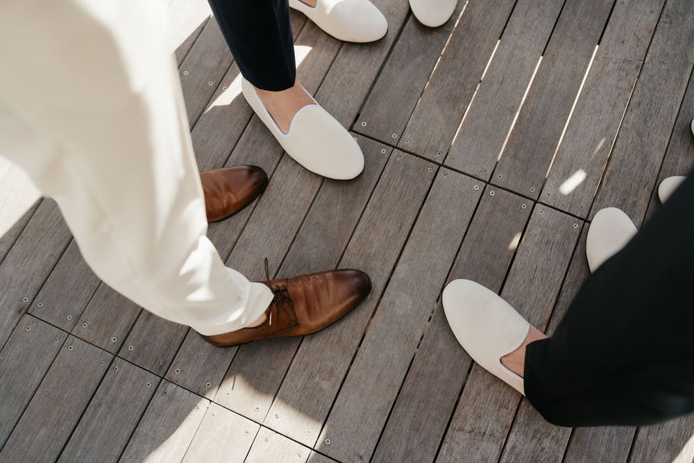 View from above of multiple pairs of feet wearing contrasting brown leather shoes and white slip-ons on wooden deck.