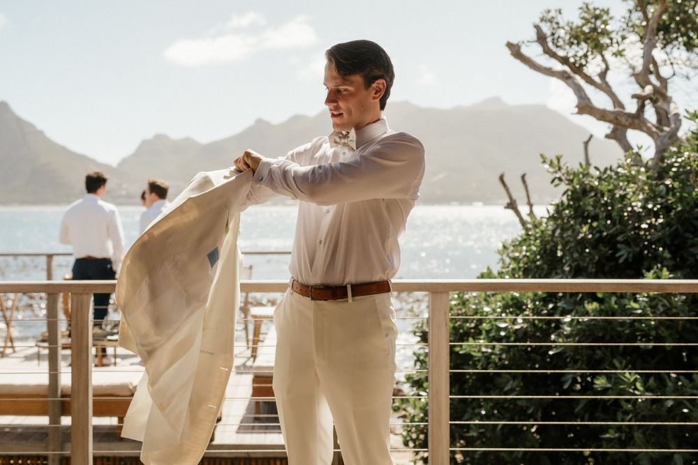 A groom in light attire adjusts his jacket with an oceanfront mountain backdrop.