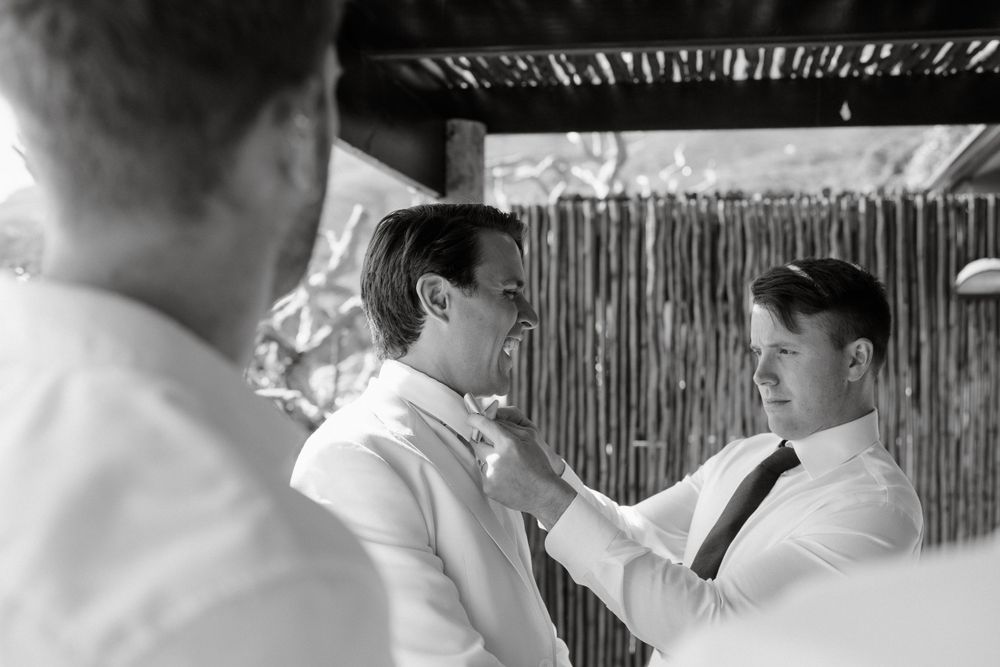 Black and white photo of two men in formal attire adjusting ties at an outdoor venue.