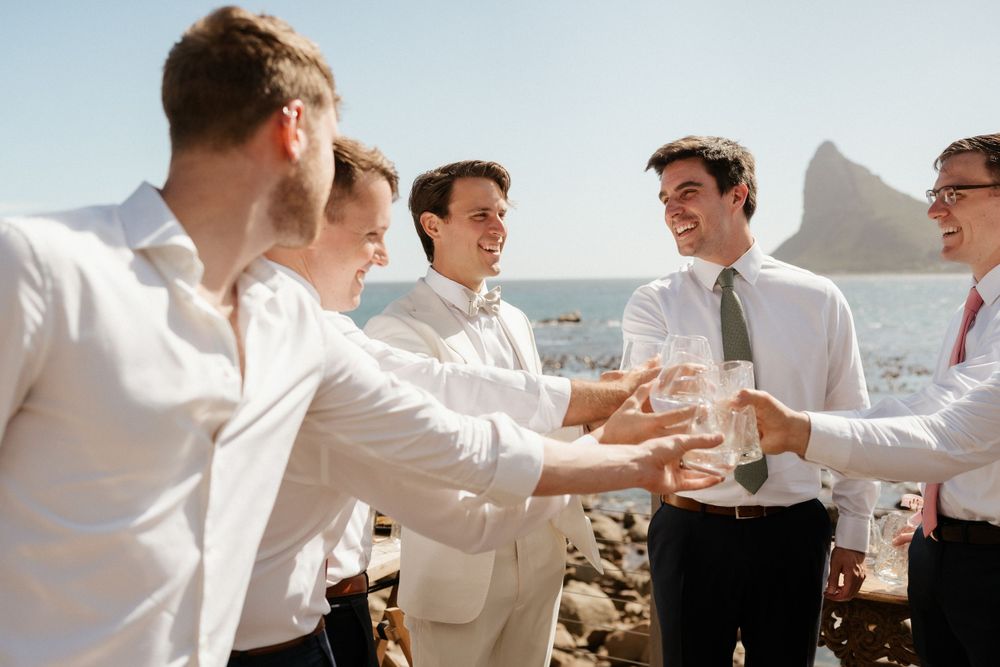 Group of groomsmen in white shirts laughing and toasting by the ocean.