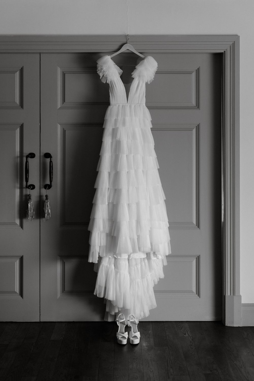 A white layered tulle wedding dress and shoes hanging on a gray door.