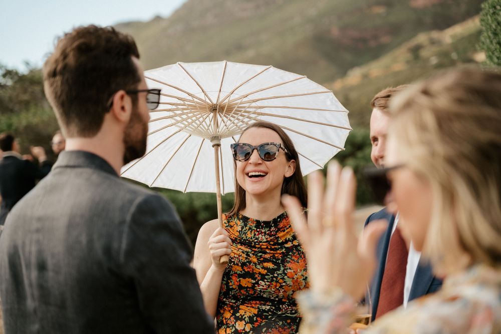 Wedding guest holding white parasol laughs with others during outdoor celebration.