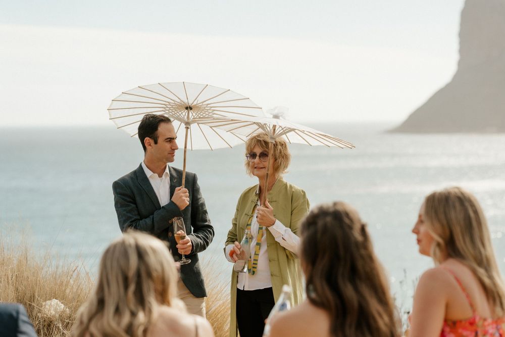 Wedding guests with decorative parasols gathered on coastal cliff with ocean view.