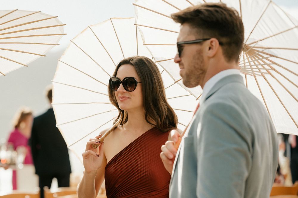 Guests with parasols enjoying an outdoor wedding celebration in elegant attire.