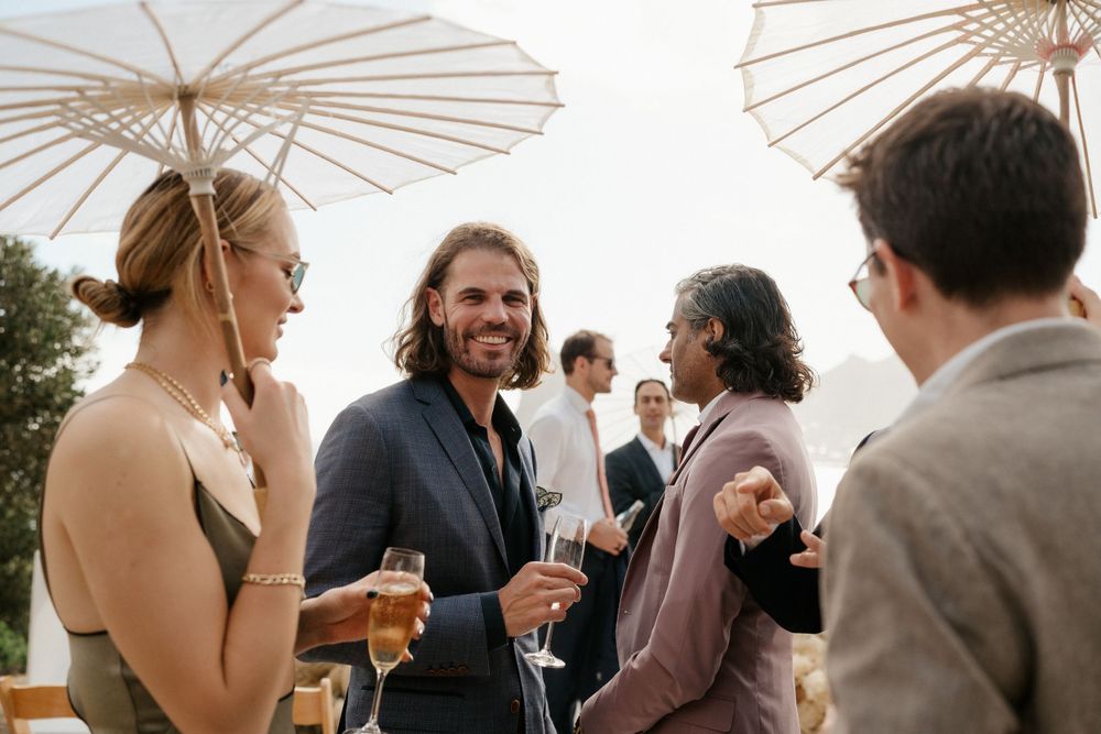 Wedding guests enjoying cocktail hour with parasols and champagne at an outdoor reception.