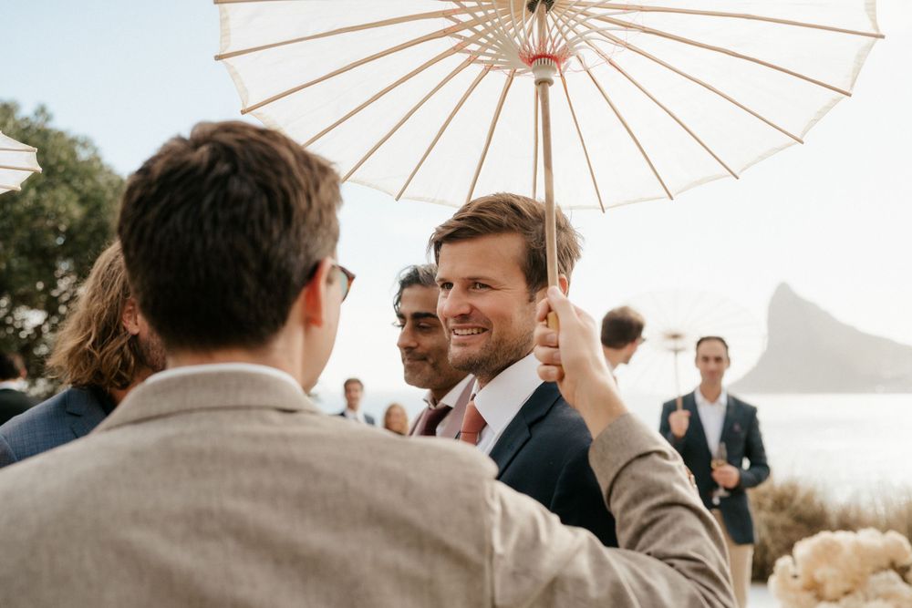 Wedding guests gathered under a white paper parasol with coastal mountains in background.