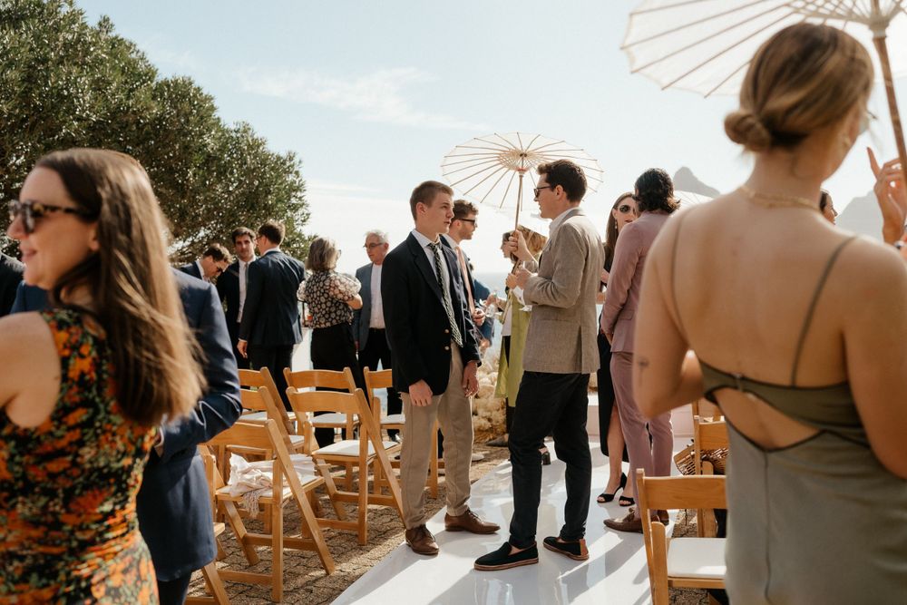 Wedding guests mingle outdoors under a white parasol during a sunny celebration.
