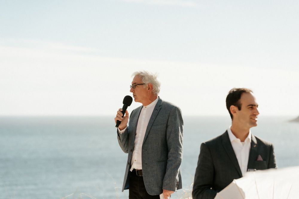 Two speakers address guests at seaside wedding ceremony.