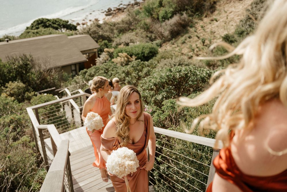 Wedding party descending coastal stairs with bouquets on a sunny day.