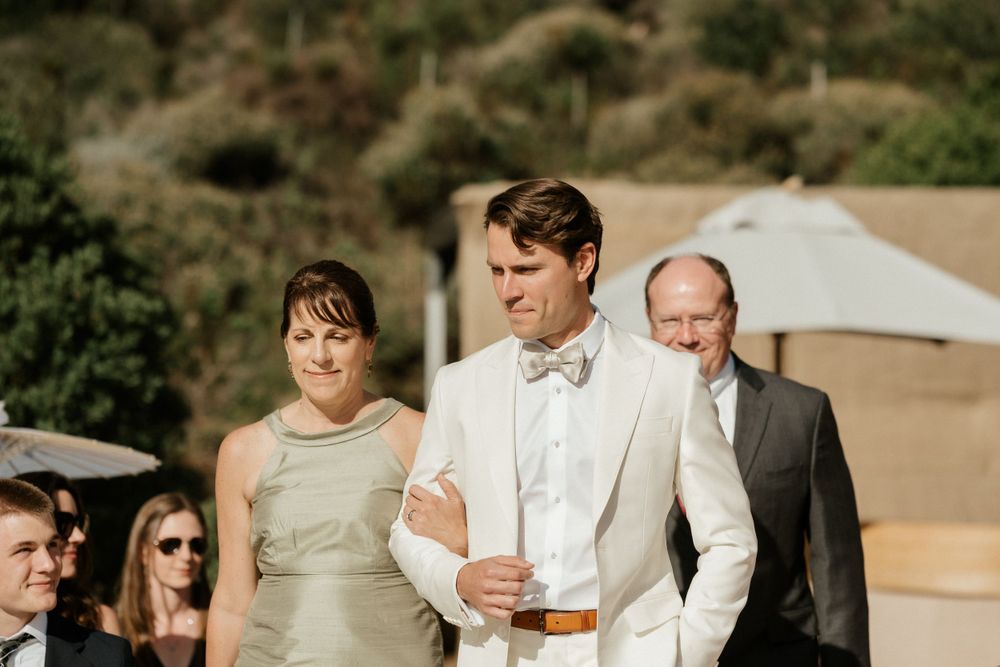 Outdoor wedding ceremony with guests seated under umbrellas against a hillside backdrop.