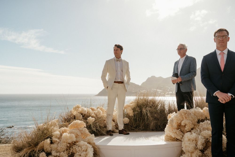 Wedding guests gather on a coastal overlook decorated with dried pampas grass arrangements against a mountainous backdrop.