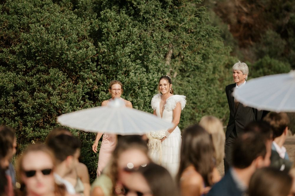Wedding ceremony guests shaded by white parasols in a sunny outdoor setting.