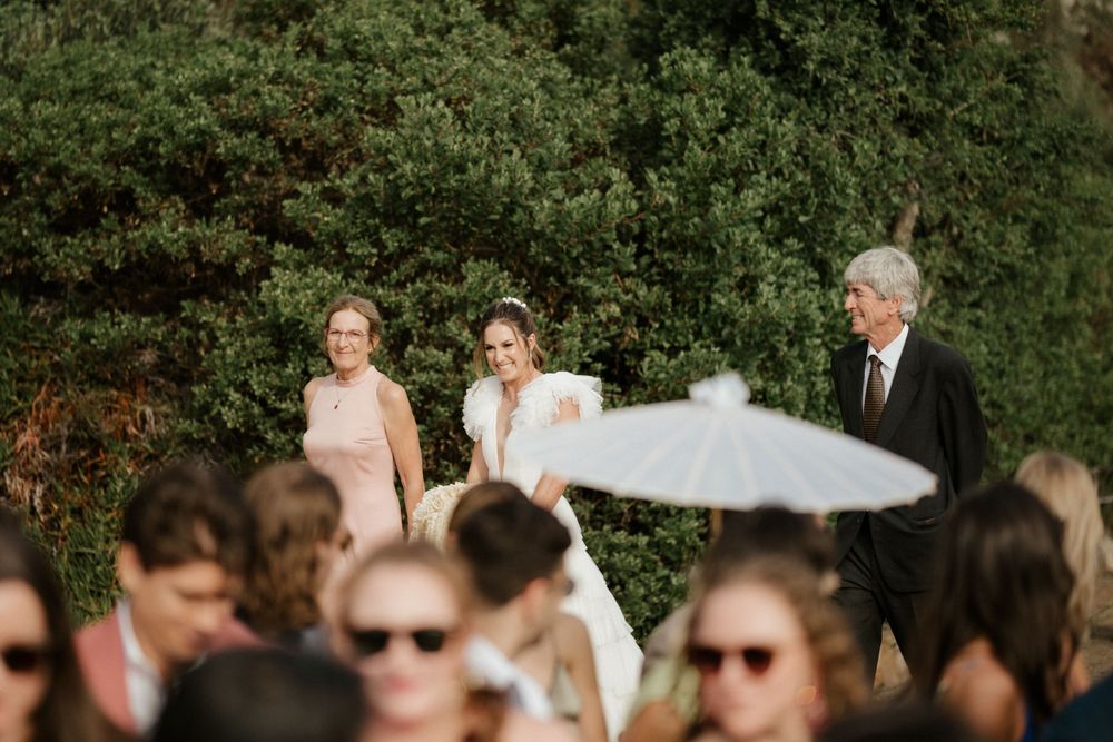 Wedding ceremony guests shielding from sun with white parasols against lush green backdrop.