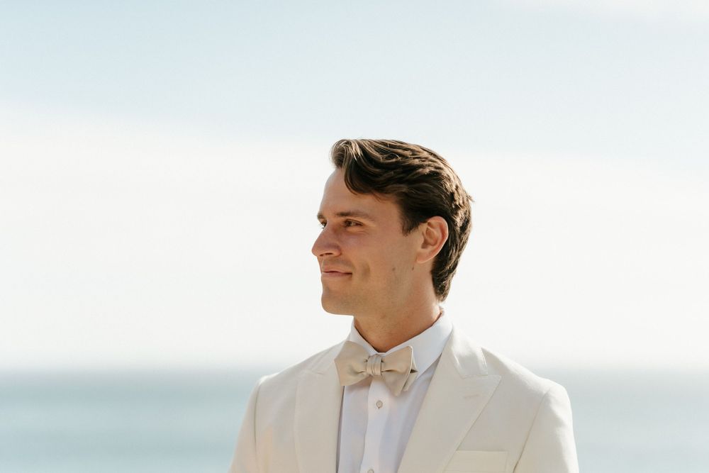 Person in white suit jacket and bow tie smiling against ocean backdrop.
