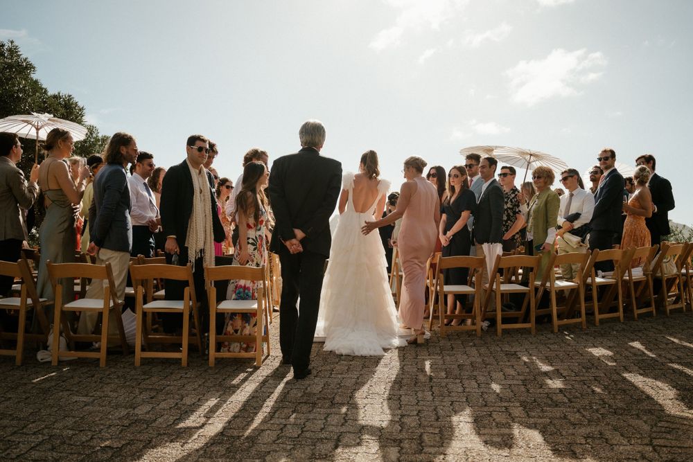 Wedding guests standing at outdoor ceremony with wooden chairs at sunset.