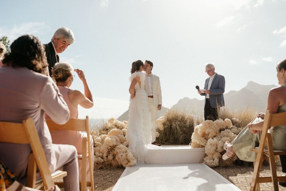 Sun-drenched coastal wedding ceremony with guests seated and mountains in the background.