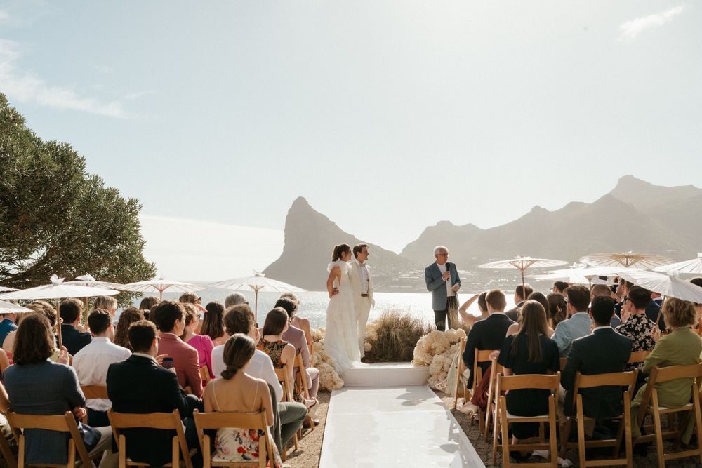 Scenic outdoor wedding ceremony with mountain backdrop and white parasols shading guests.