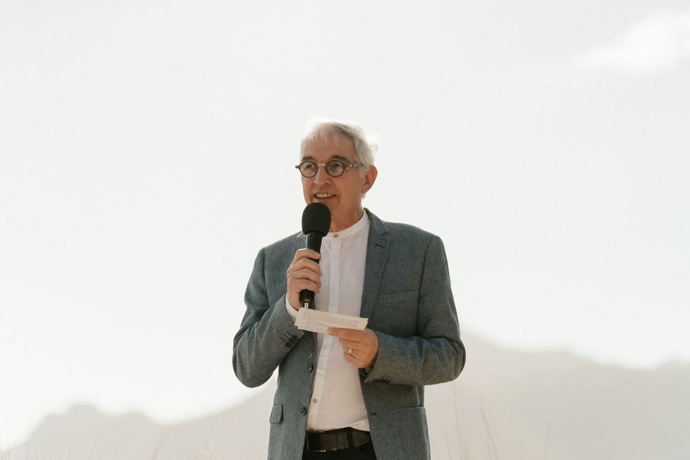 A speaker in a grey suit addresses guests at an outdoor ceremony with mountains in the background.