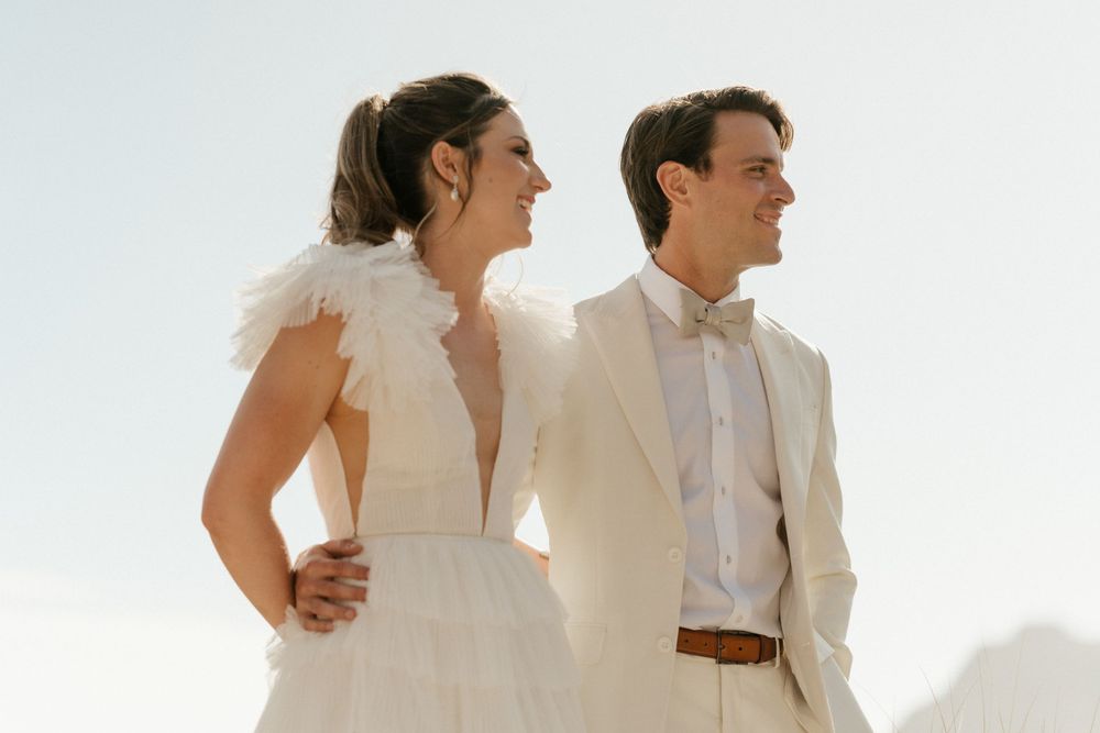 Couple in matching white outfits share joyful moment against bright sky background.
