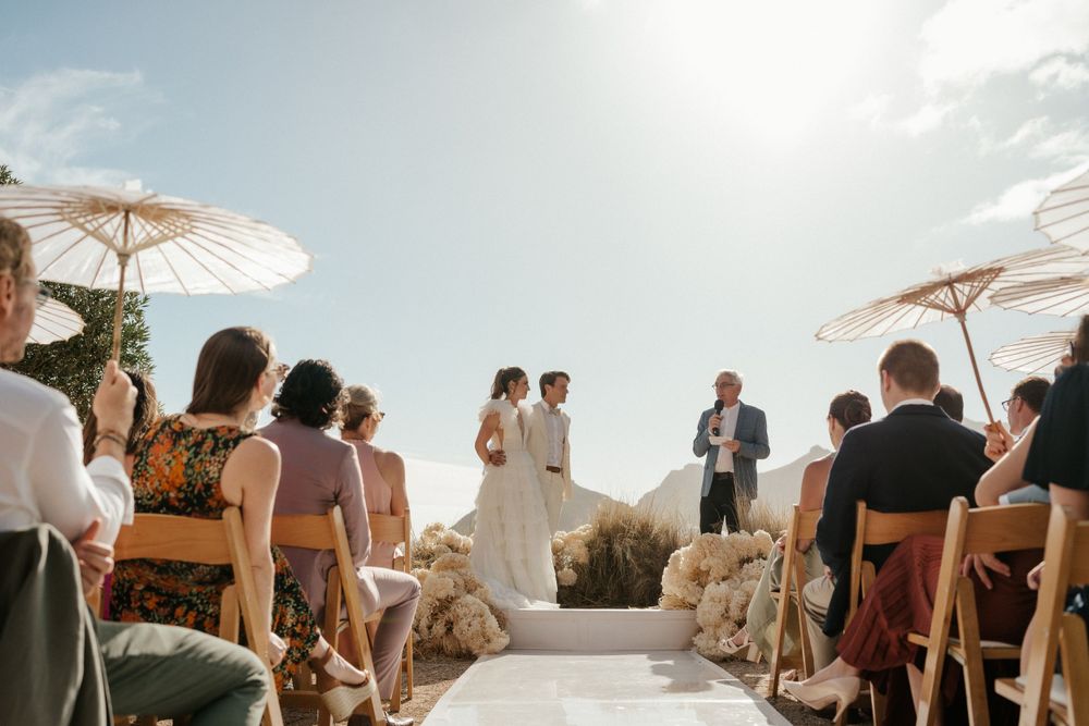 An outdoor wedding ceremony with white parasols and mountain views as guests sit on wooden chairs.