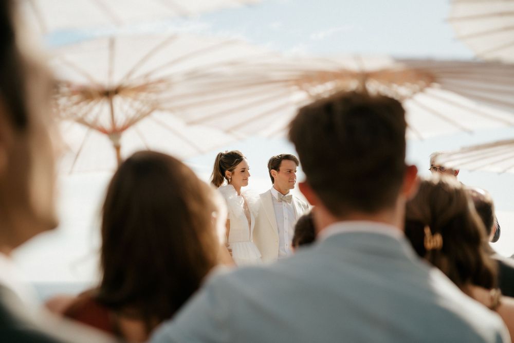 Guests gather under white parasol umbrellas at an outdoor wedding ceremony.