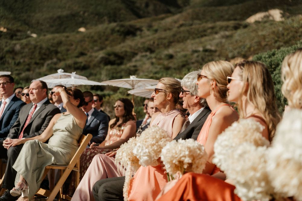Wedding guests seated in wooden chairs with parasols enjoying an outdoor ceremony.