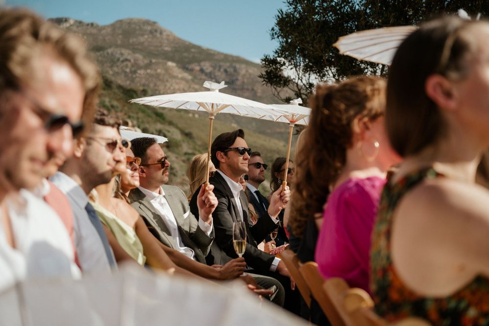 Wedding guests gathered outdoors under a sunny sky with mountains in the background.