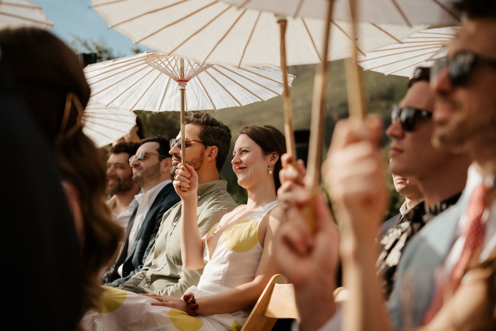 Wedding guests sitting under white paper parasols during an outdoor ceremony.