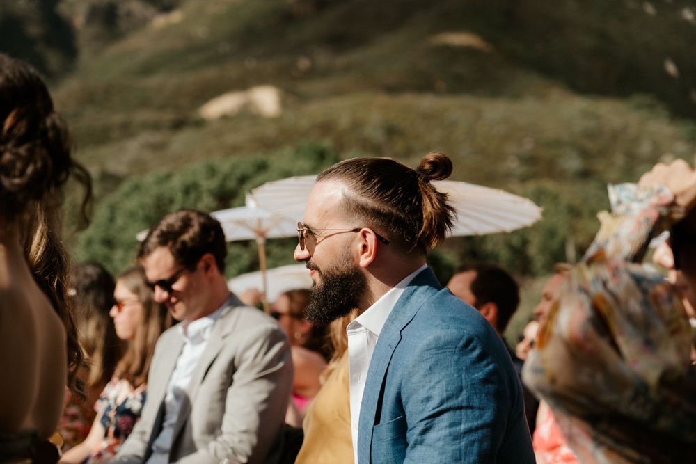 Wedding guests gathered outdoors with mountains in background and white parasols for shade.