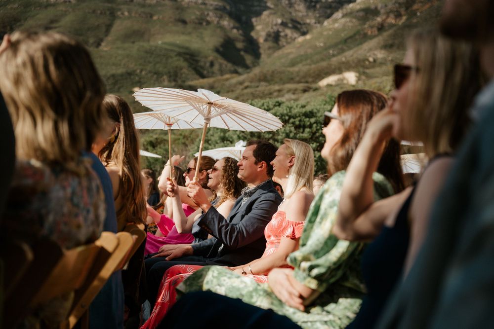 Wedding guests seated with parasols on a mountainside during an outdoor ceremony.
