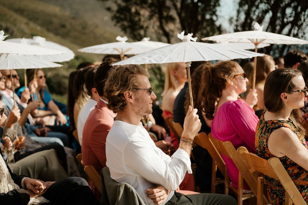 Wedding guests seated with white parasols at an outdoor ceremony in the mountains.