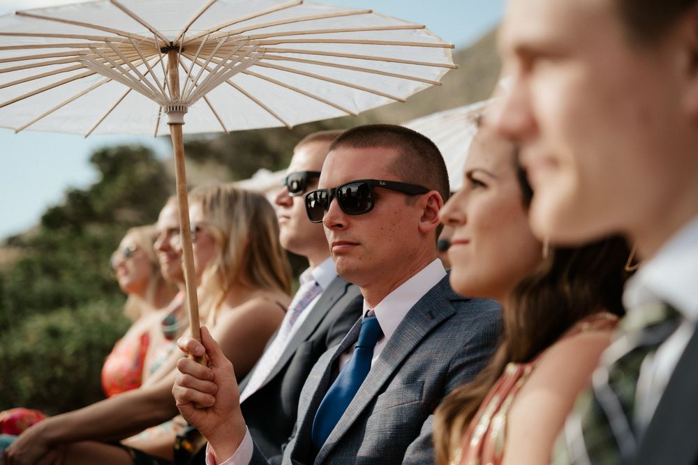 Wedding guests sit in chairs outdoors with decorative parasols providing shade.