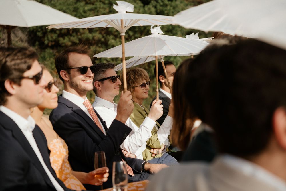 Wedding guests enjoy drinks under white umbrellas at an outdoor reception.