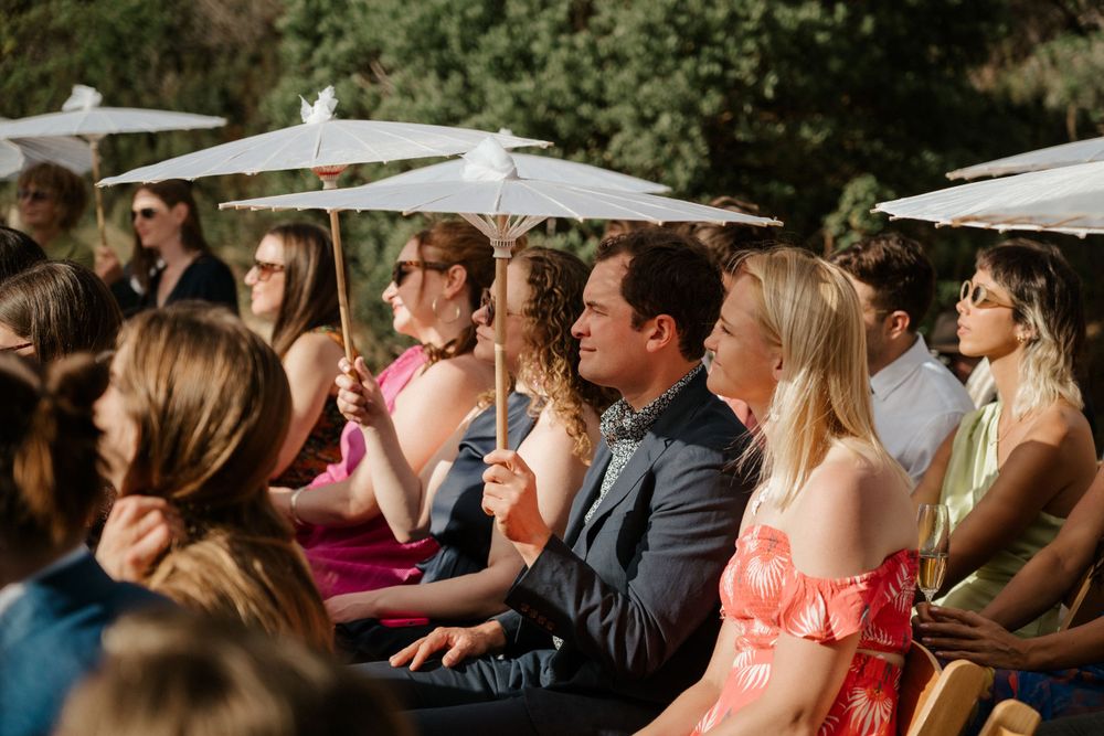 Wedding guests sit beneath white parasols during an outdoor ceremony on a sunny day.