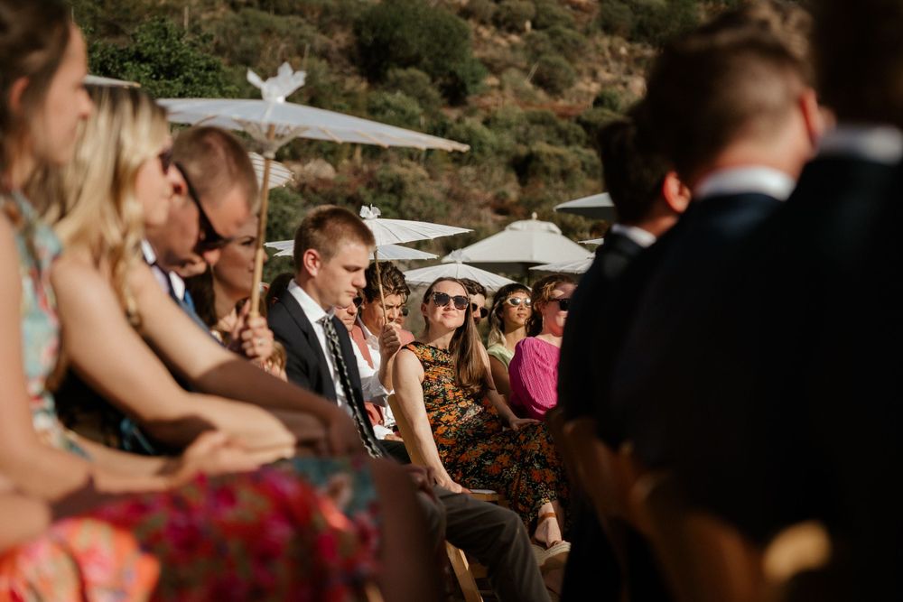 Wedding guests seated outdoors at a sunny mountainside ceremony with white umbrellas providing shade.