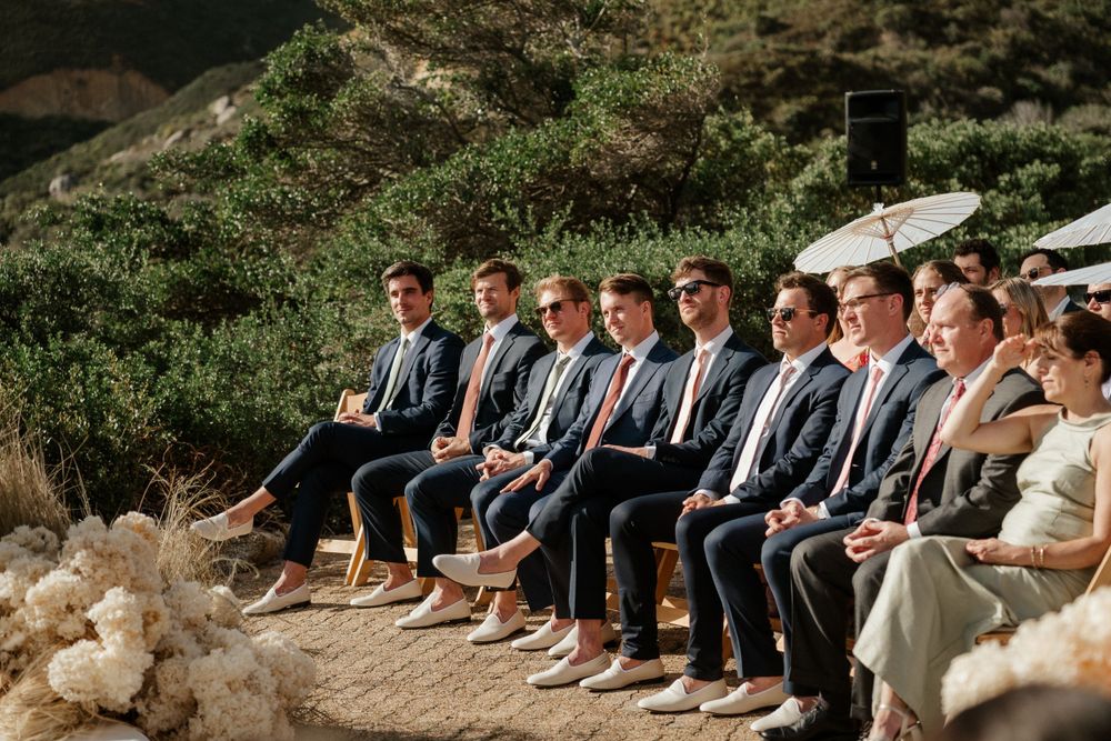 Wedding party members in suits sit together on stone steps against a mountainous backdrop.